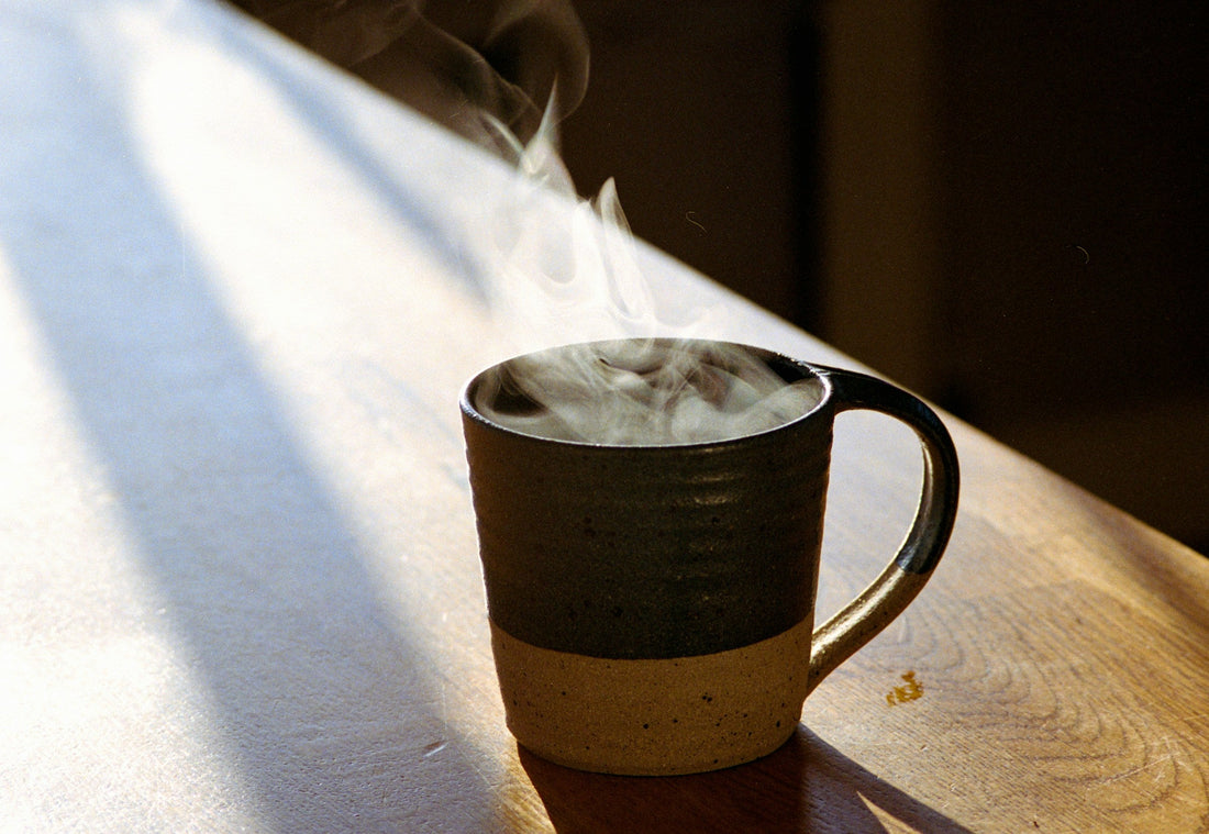 Steam rises from a stoneware mug catching low light on a wooden table